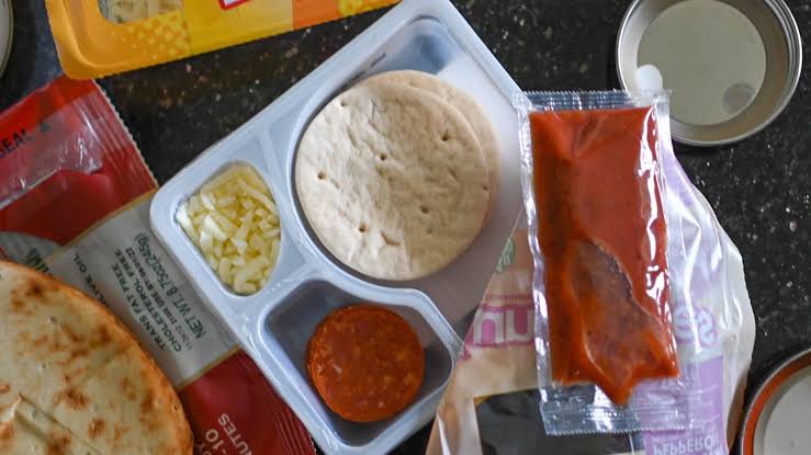 A lunch kit tray with mini pizza crusts, a compartment of shredded cheese, pepperoni, and a packet of pizza sauce, surrounded by packaging on a dark surface.