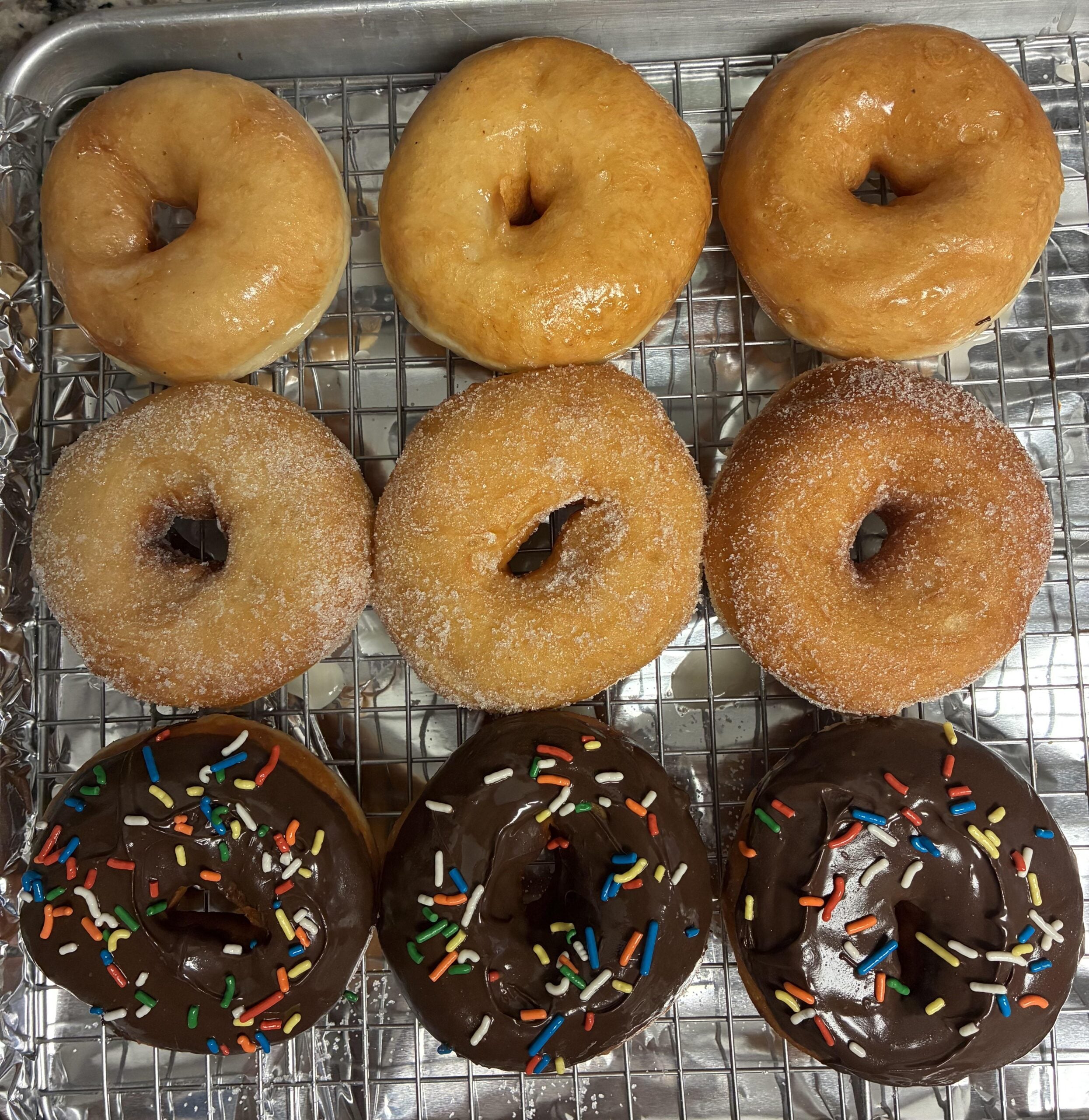 Nine donuts on a cooling rack: three plain glazed, three sugar-coated, and three chocolate-glazed with colorful sprinkles, all arranged in neat rows.