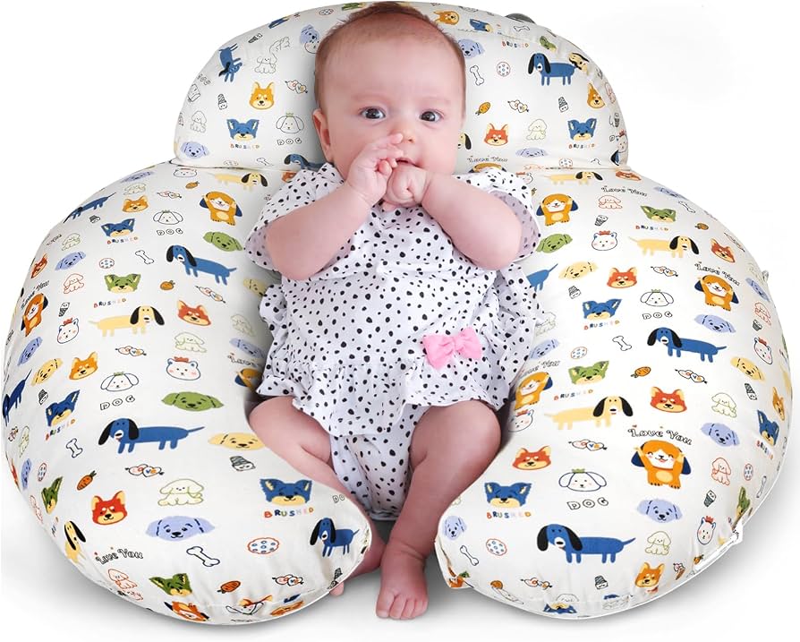 A baby in a polka dot outfit sits in a U-shaped pillow with colorful cartoon animals and bones printed on it.