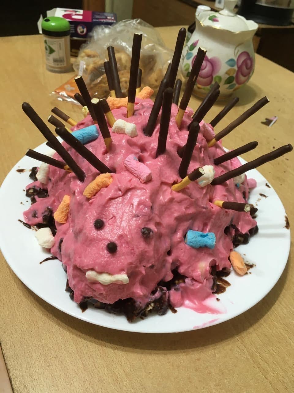 A hedgehog-shaped cake with pink frosting, chocolate sticks as spikes, and colorful marshmallows on top, sitting on a white plate on a wooden table.