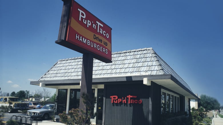 A retro Pup ‘n’ Taco fast food restaurant with a large red sign reading “Pup ‘n’ Taco Drive Up Hamburgers” stands under a clear blue sky. Several cars are parked nearby.