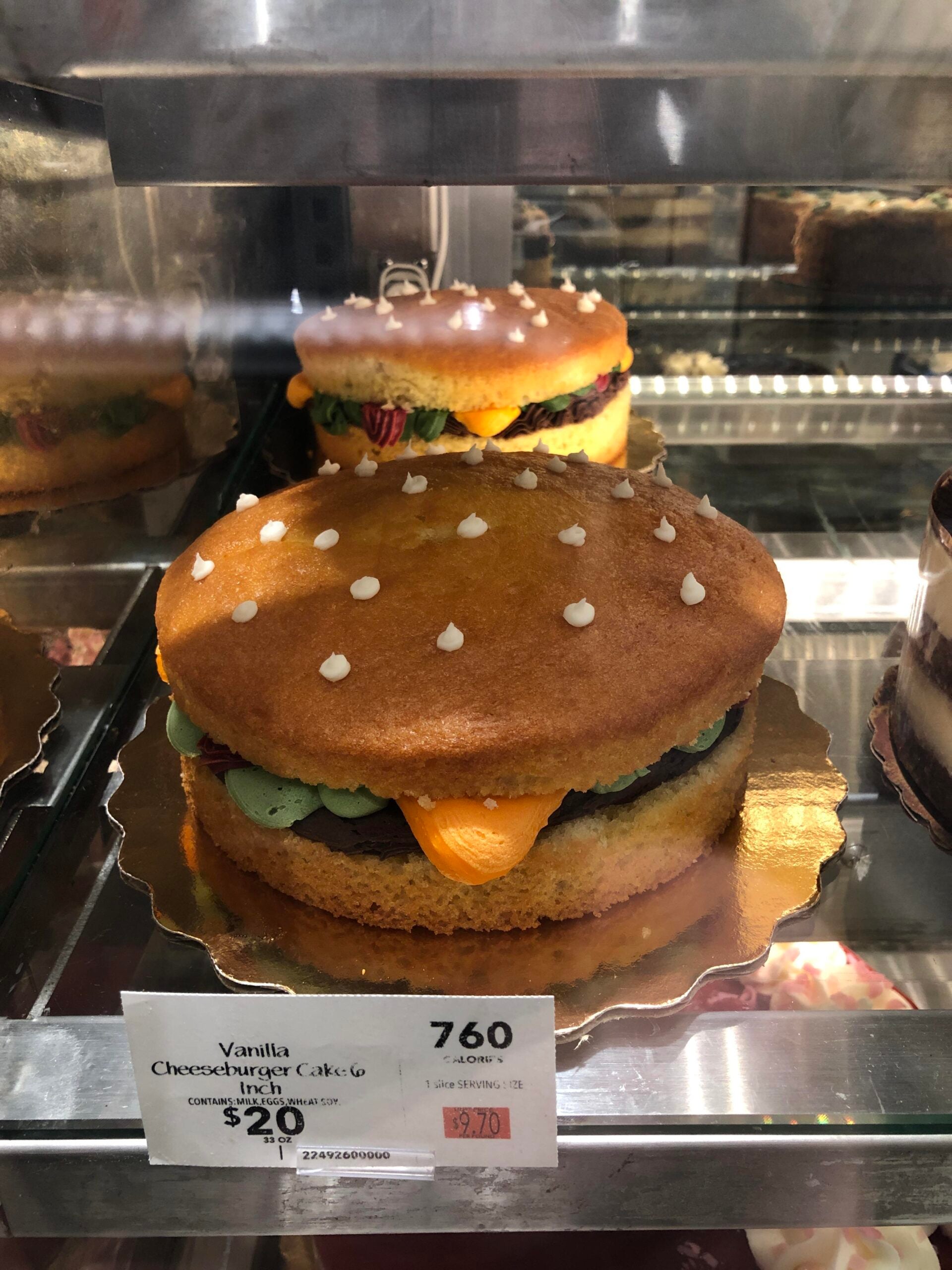 A cake decorated to look like a cheeseburger, with layers resembling a bun, lettuce, and cheese. It’s displayed in a bakery case with a price tag of $20 in front.