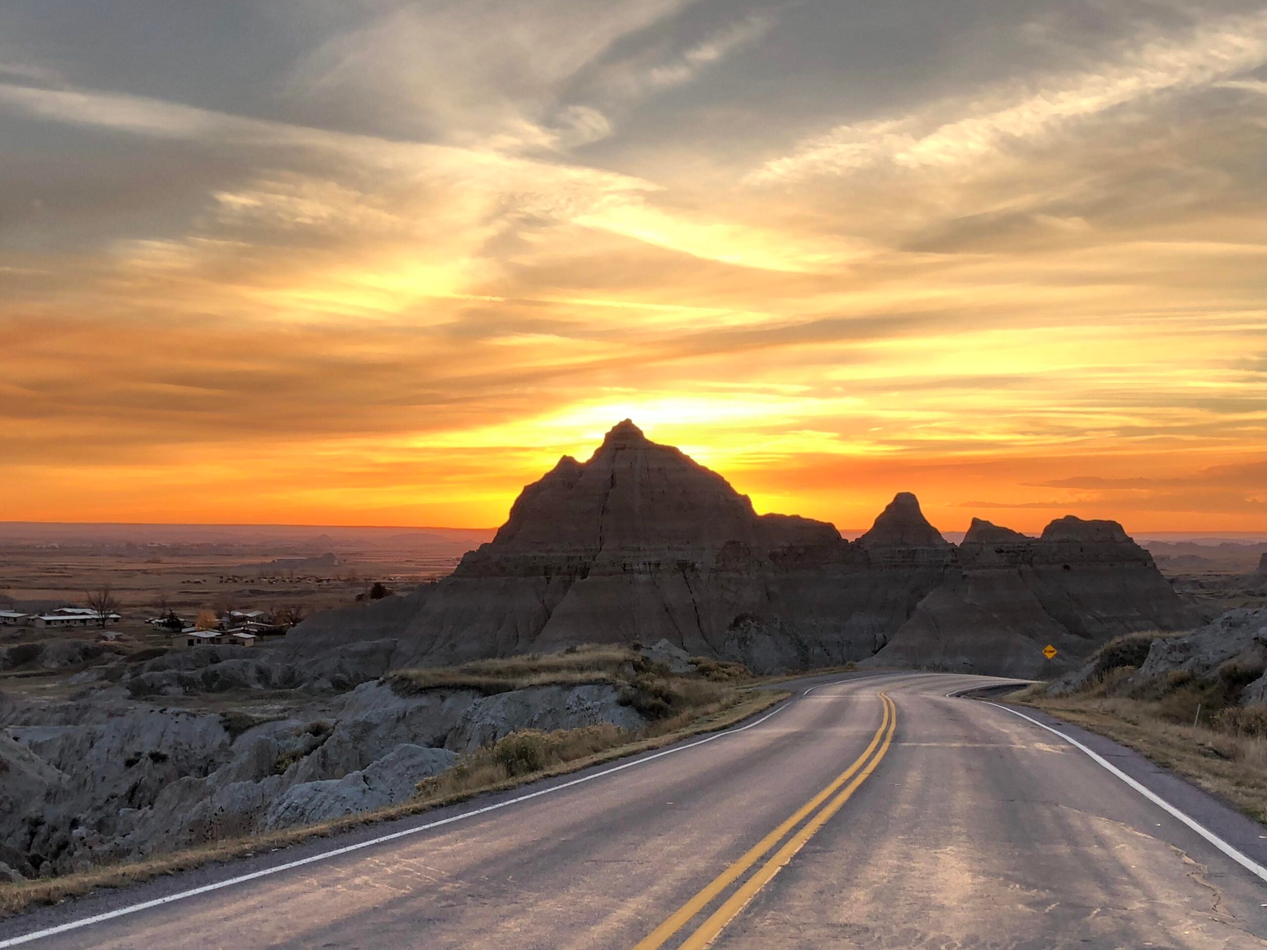 A winding road leads toward rocky hills under a dramatic sunset sky, with golden and orange hues lighting up clouds above the landscape—an inviting scene that perfectly captures the spirit of memorable road trips.
