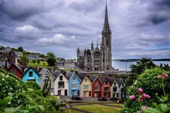 Colorful houses line a hillside in front of a large stone church with a tall spire, overlooking a body of water under a cloudy sky. Lush greenery and flowers frame the foreground.