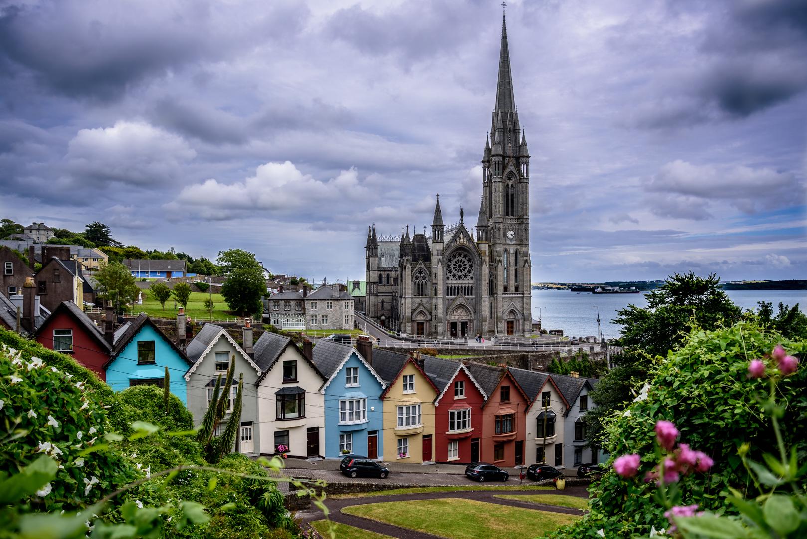 Colorful houses line a hillside in front of a large stone church with a tall spire, overlooking a body of water under a cloudy sky. Lush greenery and flowers frame the foreground.