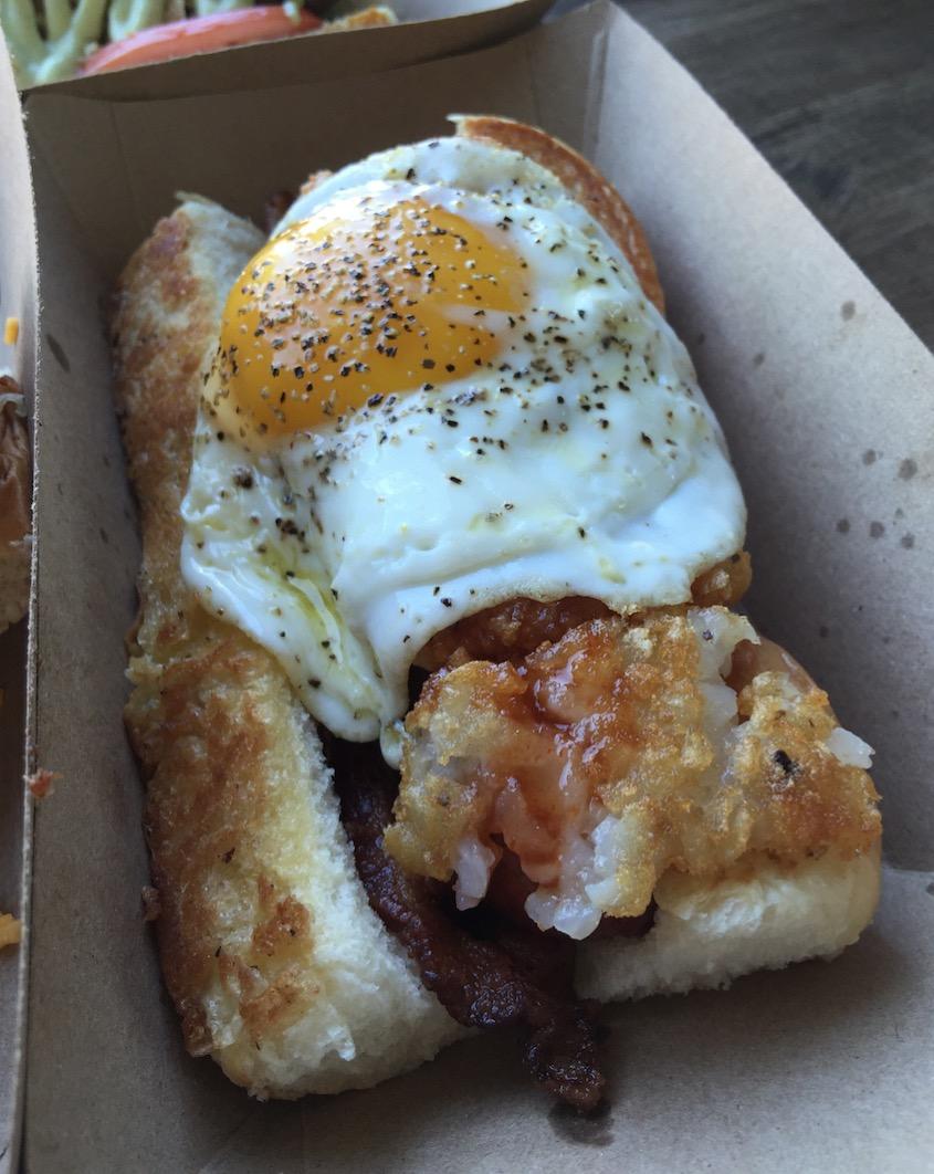 A sandwich in a cardboard tray topped with crispy bacon, a golden fried egg, and a battered fried item on a toasted bun.