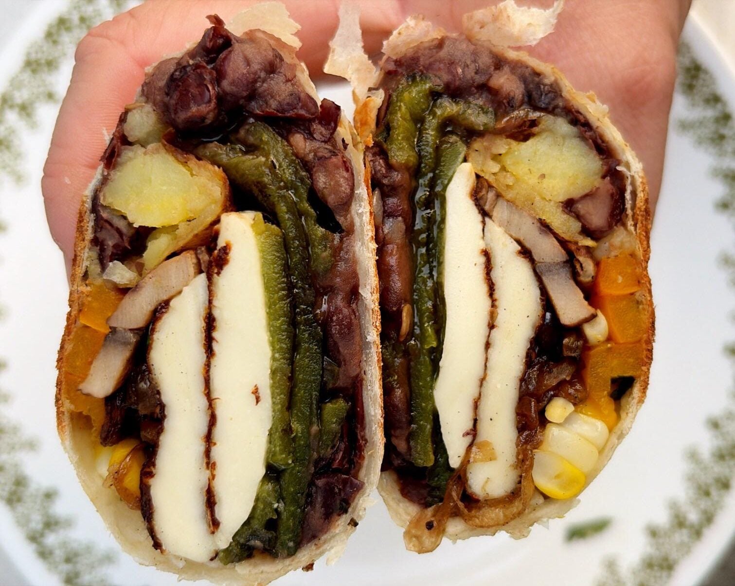 A hand holds a split burrito showing layers of beans, cheese, potatoes, roasted peppers, mushrooms, corn, and other vegetables on a patterned plate.