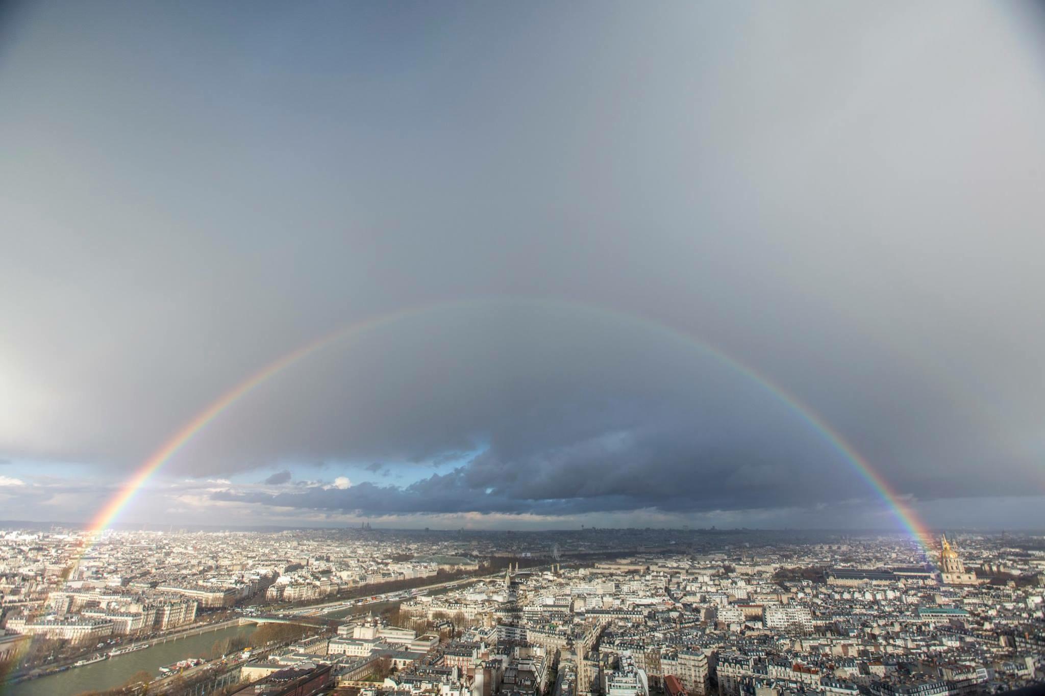 A wide rainbow arcs over the cityscape of Paris, with the Seine River running through the center and clouds filling the sky.
