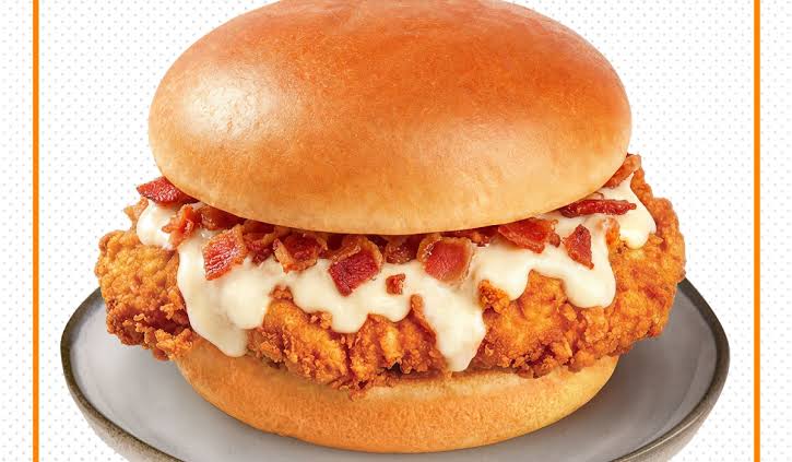 A crispy fried chicken sandwich topped with white sauce and bacon bits, served on a soft, shiny bun, placed on a gray plate.