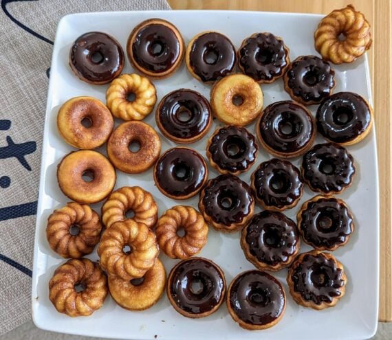 A square plate holds mini donuts&mdash;some plain, some ring-shaped, and others topped with shiny chocolate glaze&mdash;in neat rows. The donuts are golden brown and neatly arranged.