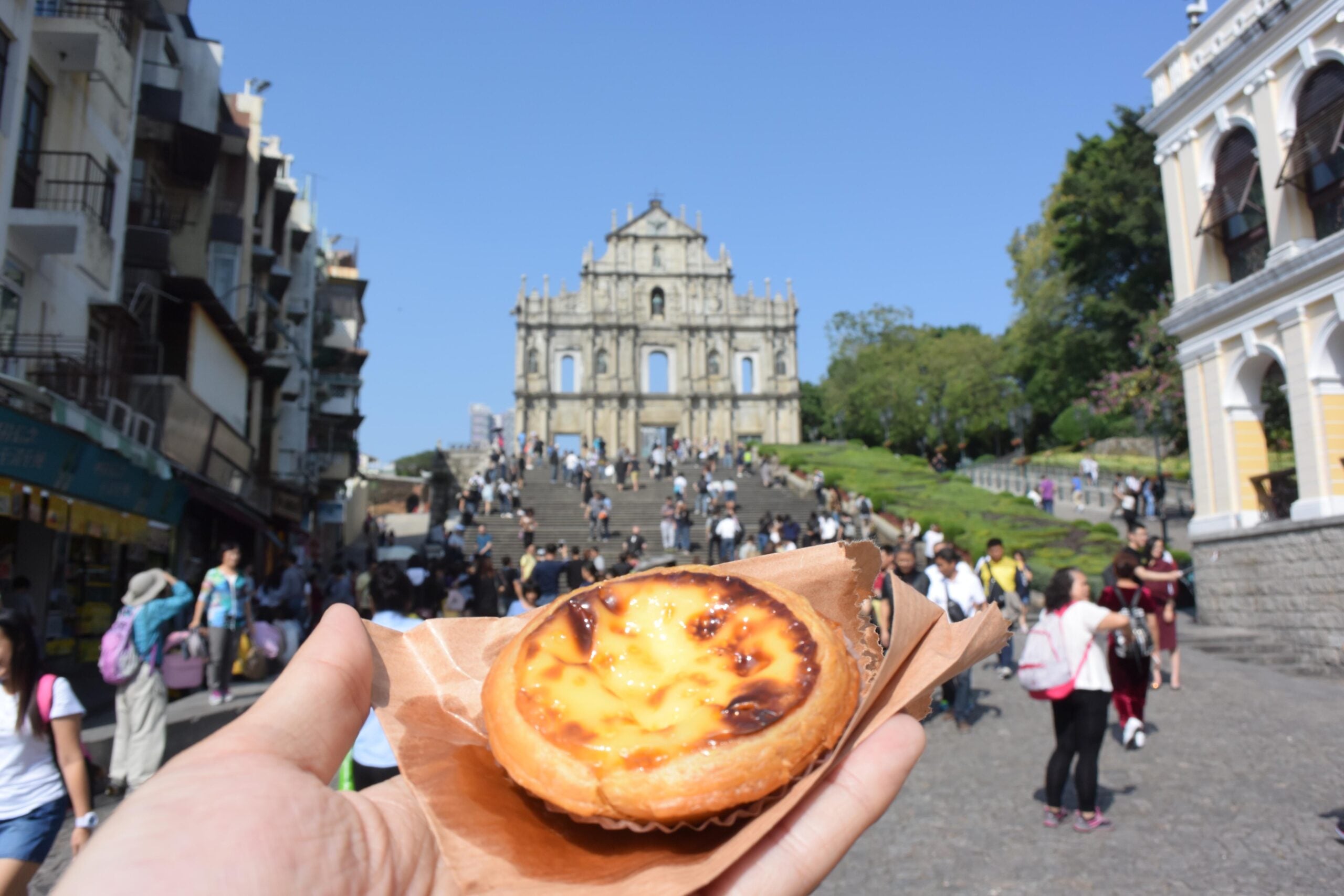 A hand holds an egg tart in the foreground, showcasing one of the amazing food people ate while on vacation, with the Ruins of St. Paul’s in Macau and a busy crowd on the steps in the background on a sunny day.