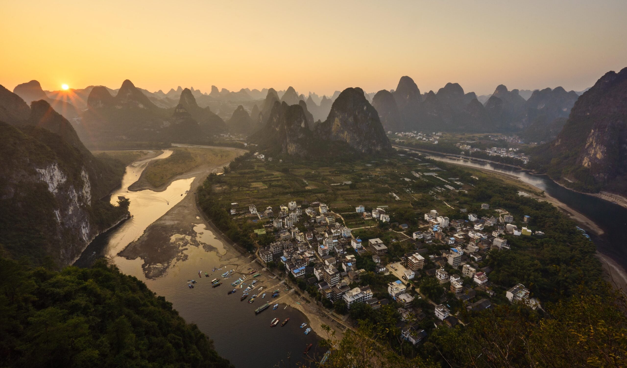 A river bends around a village with boats, surrounded by dramatic, steep karst mountains at sunset. The sky glows orange, and lush greenery covers the landscape.