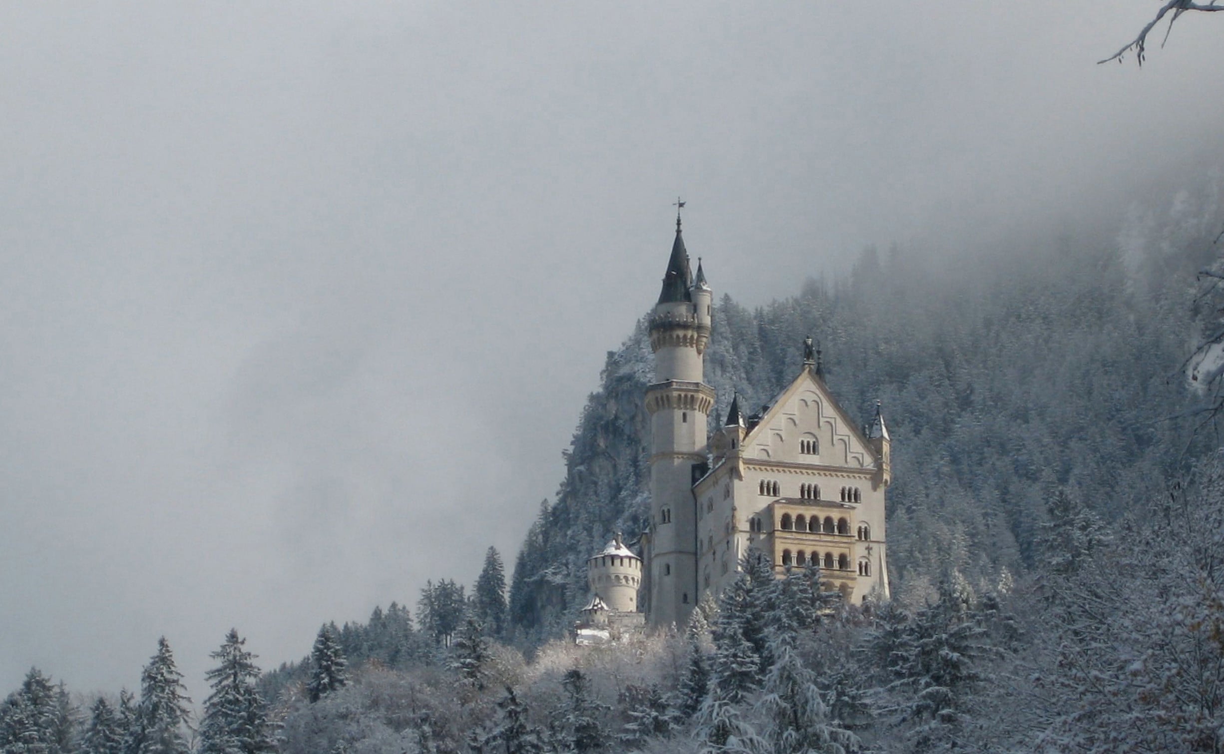 A fairytale-like castle sits atop a snowy hill, surrounded by snow-covered trees and misty mountains, creating a serene winter scene that invites wonder into every travelers’ journeys.