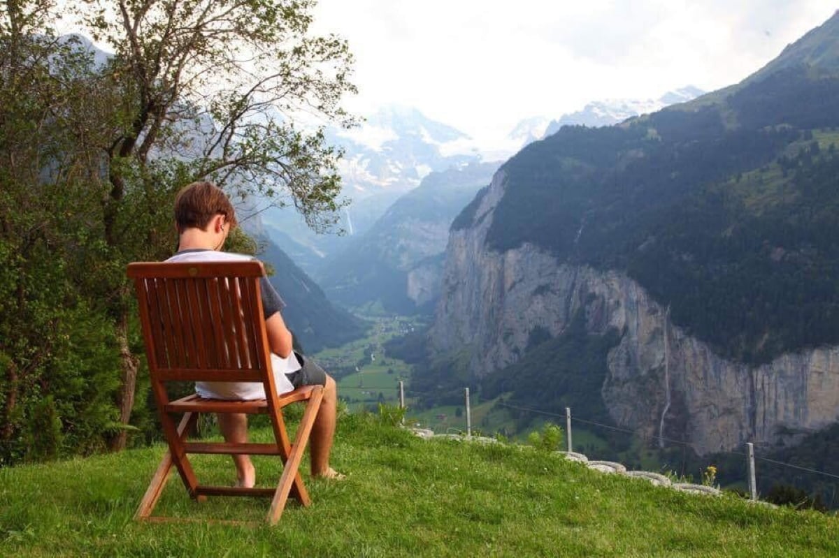A person sits on a wooden chair on a grassy hill, looking at a deep valley with tall mountains and cliffs in the background under a partly cloudy sky.