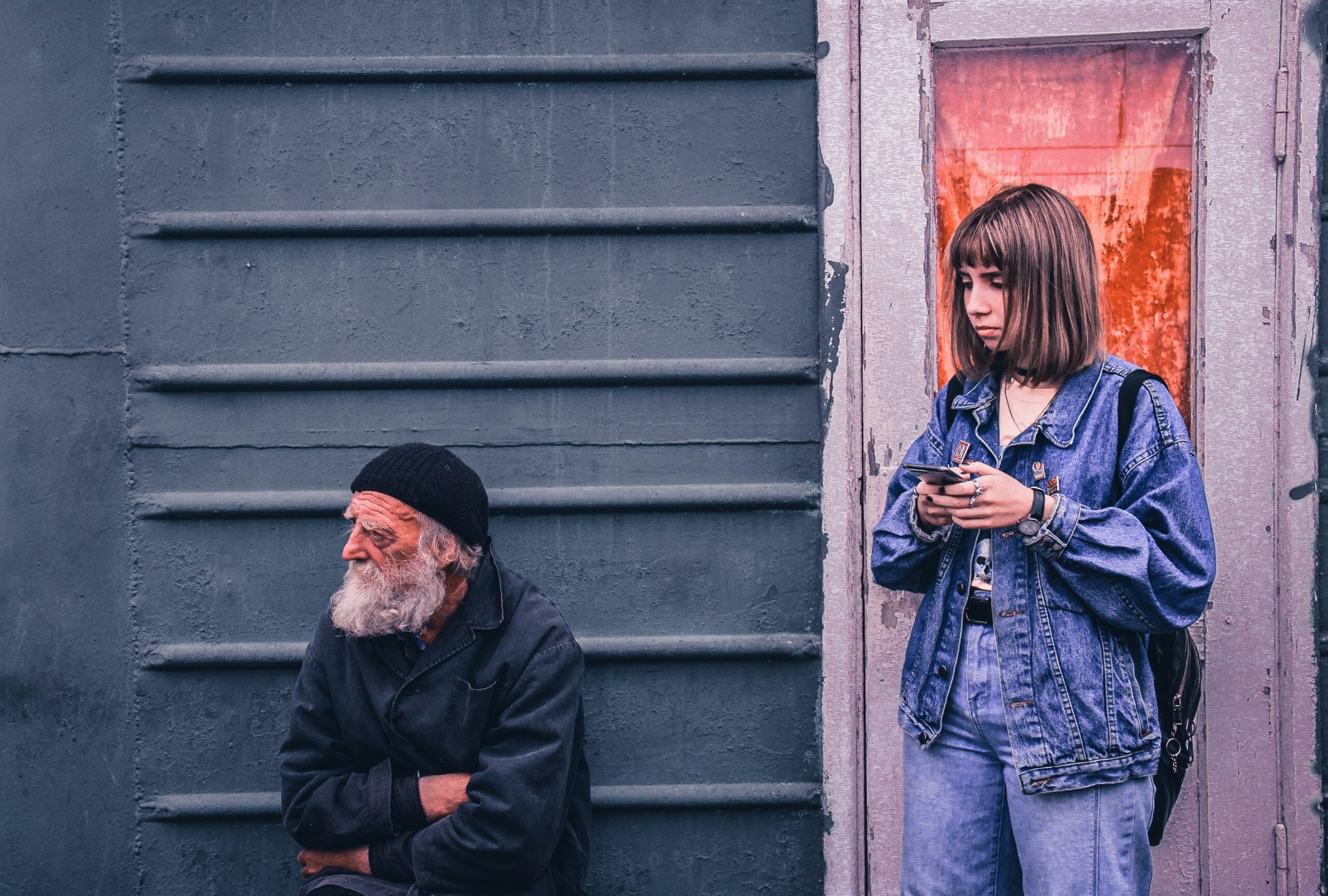 An older man with a white beard sits by a wall while a young woman in denim stands nearby, holding a phone and looking away—an everyday moment that quietly captures life in 2026. The background is gray, with the doorway lit by a reddish light.