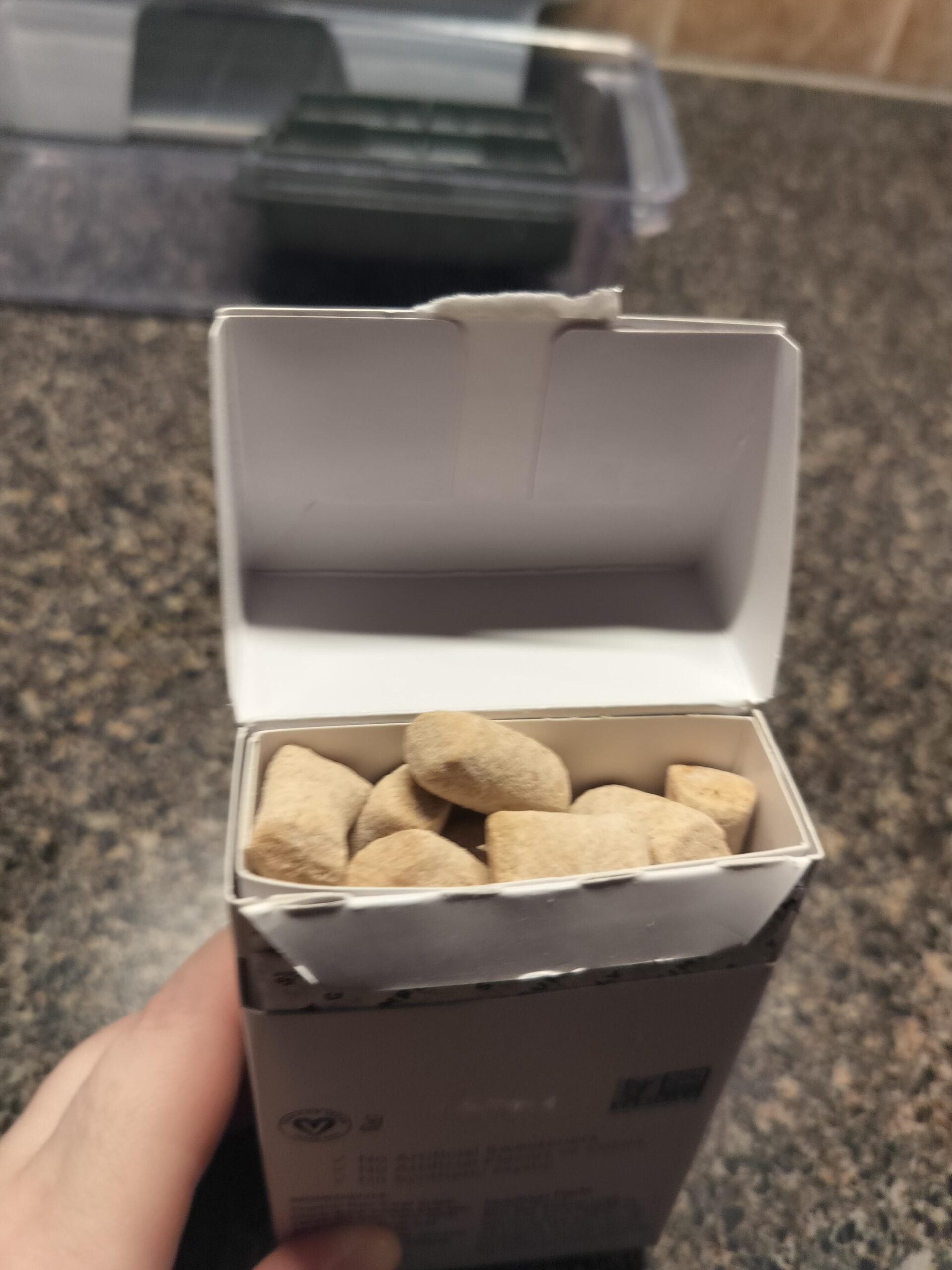 A hand holds an open white box containing several light brown, irregularly shaped chewable tablets on a dark countertop.