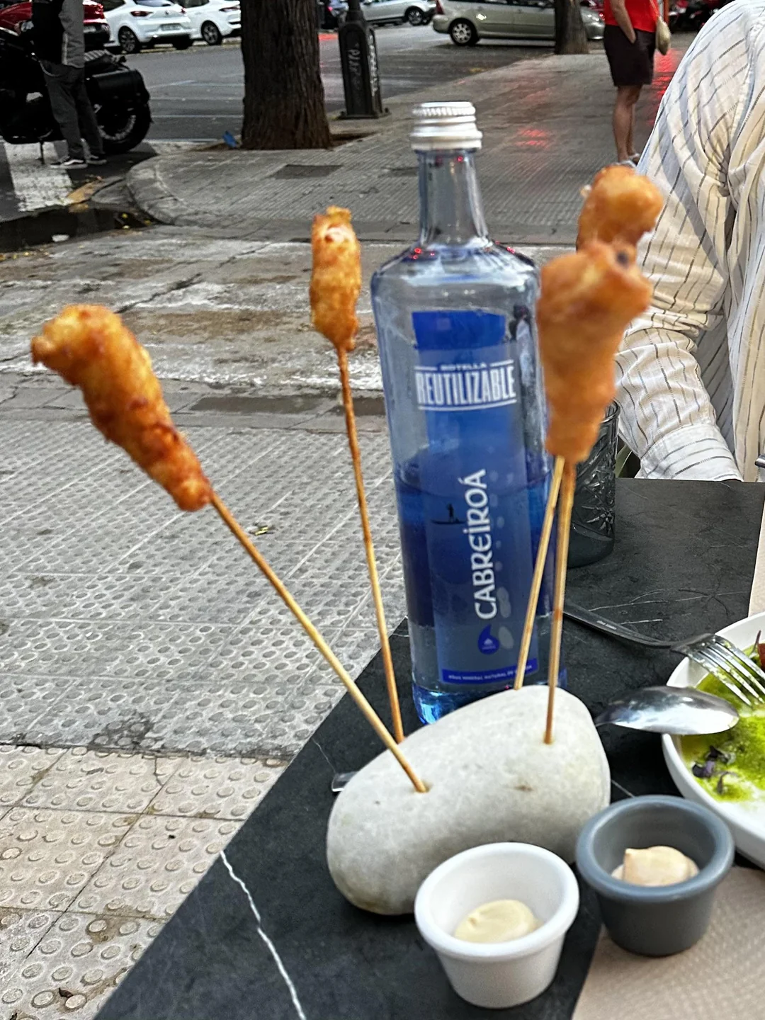 Four skewered fried foods are served upright in a white stone, with a blue water bottle and dipping sauces on a table. The scene is outdoors on a city sidewalk. Ridiculous food plating.