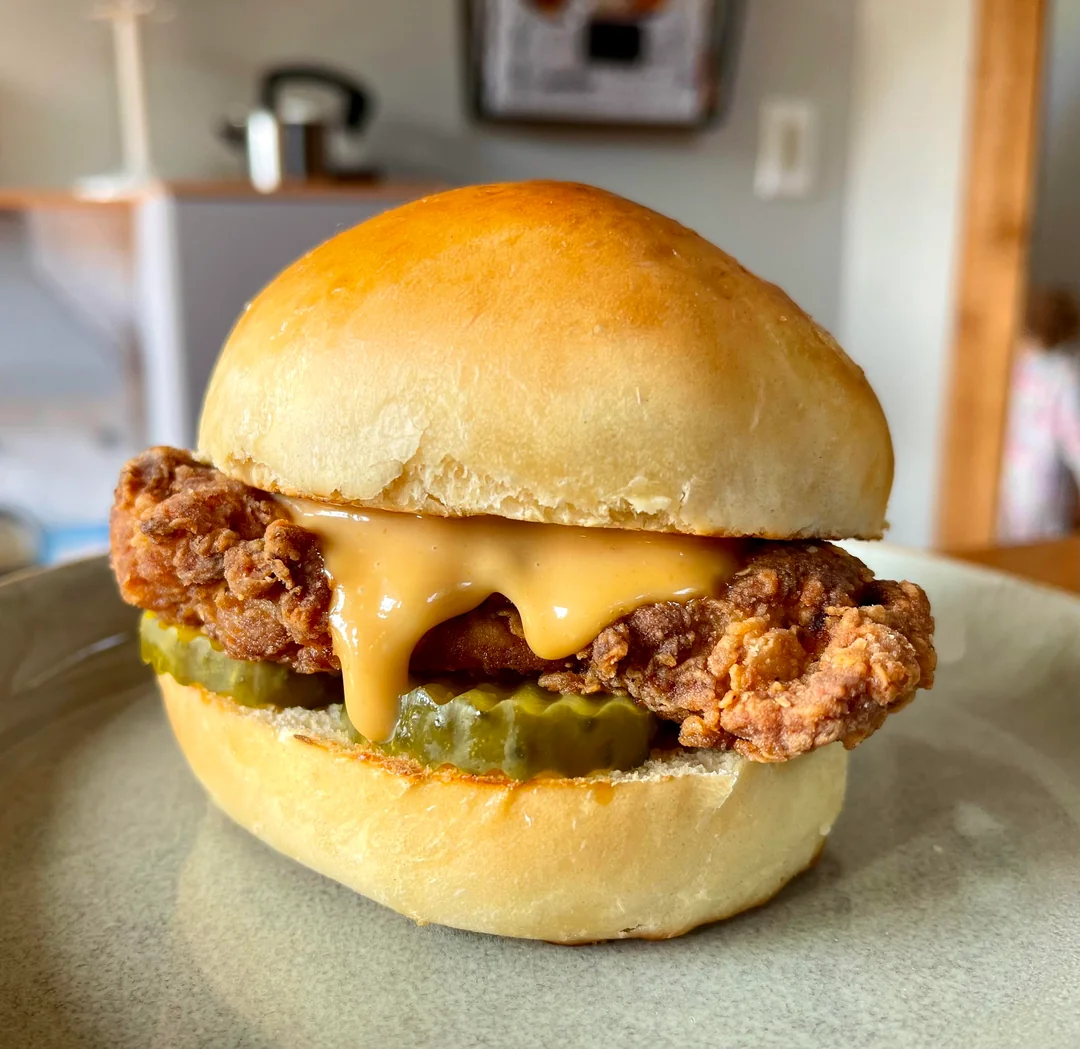 A fried chicken sandwich with melted cheese and pickles on a shiny bun, placed on a gray plate.