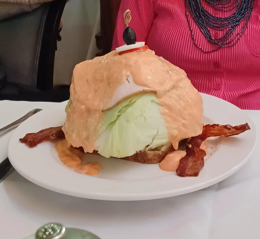 A wedge of iceberg lettuce topped with creamy orange dressing, served on toast with bacon strips on a white plate. A garnish of olive and onion is pinned on top.