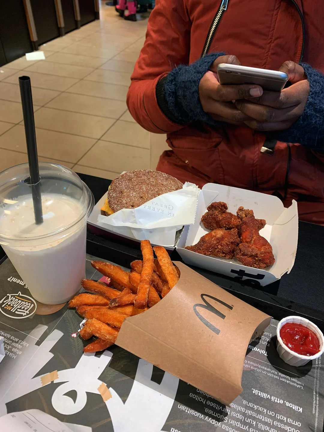 A person in a red jacket uses a phone at a table with a burger, chicken wings, sweet potato fries, ketchup, and a cold drink with a straw inside a fast food restaurant.