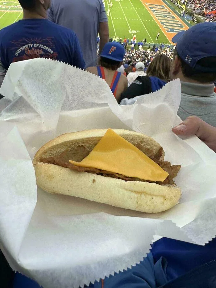 A person holds a sandwich with a burger patty and a single slice of cheddar cheese at a sports stadium, with a football game and spectators visible in the background.
