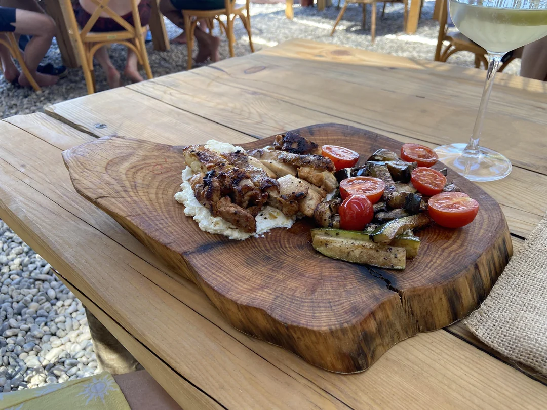 Grilled chicken, roasted zucchini, and halved cherry tomatoes on a wooden serving board atop a rustic table, with a glass of white wine nearby and people seated in the background.