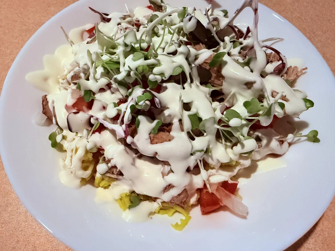 A white plate of salad topped with greens, chopped tomatoes, shredded chicken, and drizzled generously with creamy white dressing.