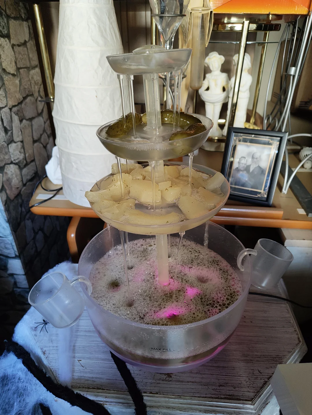 A three-tiered tabletop beverage fountain with liquid flowing down each level, plastic cups on the sides, and a pink glow at the base. The fountain is set among home decor and framed photos.