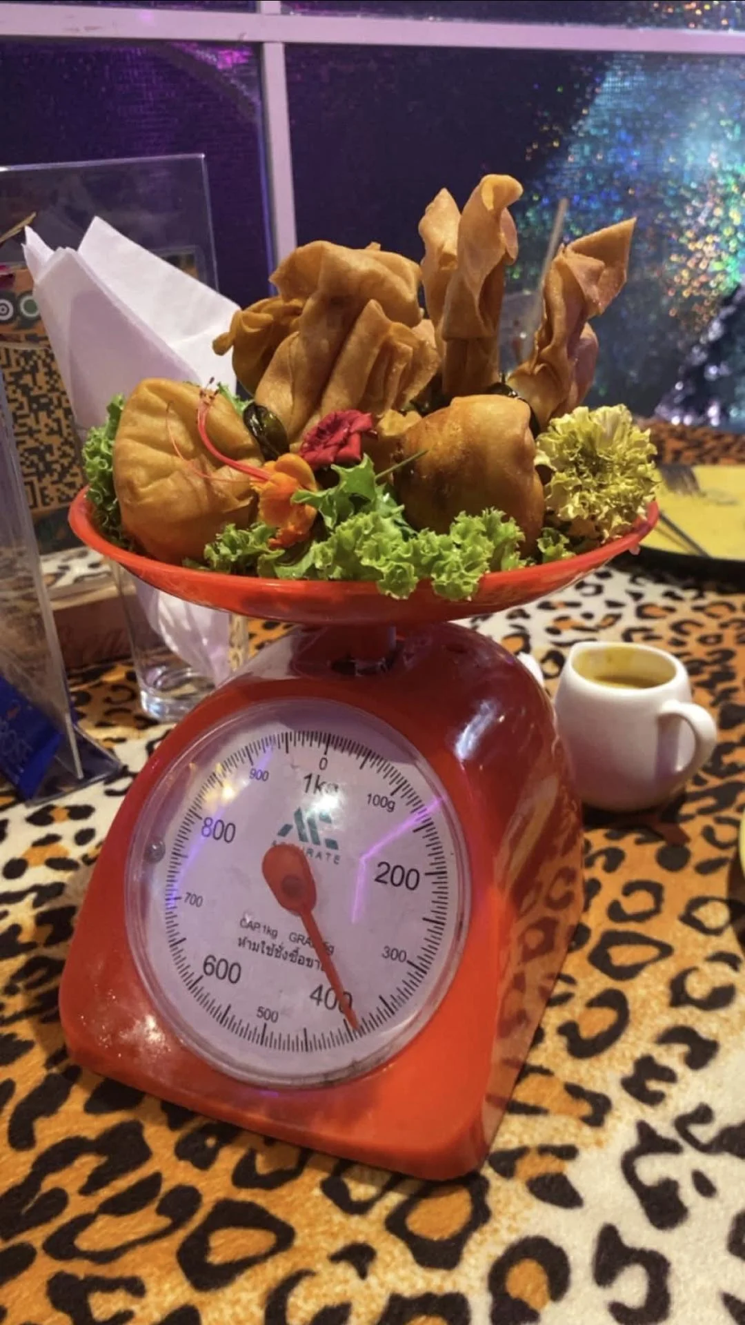 A red kitchen scale holds fried snacks and greens on top. The scale reads about 500 grams. There’s a small cup of sauce and a leopard-print tablecloth beneath. Napkins and a menu are visible in the background.