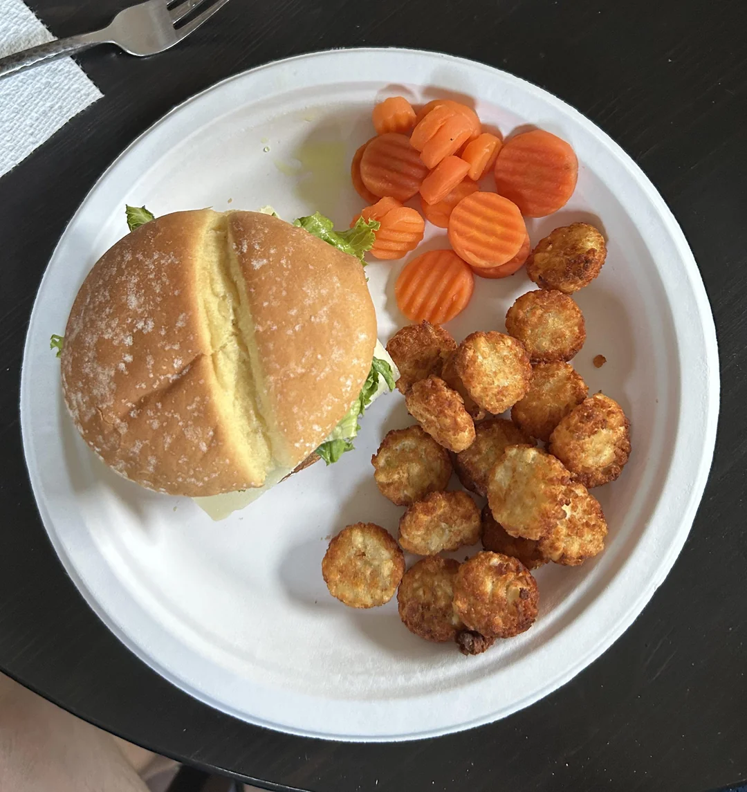 A paper plate with a sandwich on a bun, several round tater tots, and sliced cooked carrots on a dark table with a fork and napkin nearby.