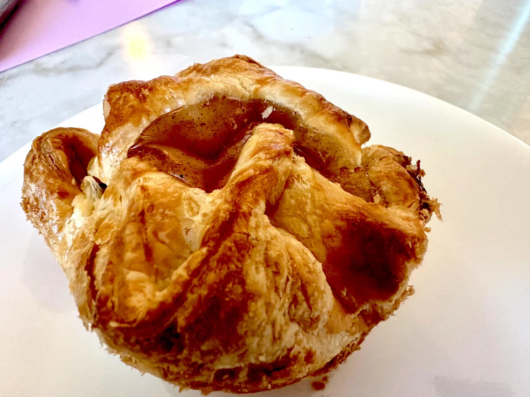 A close-up of a golden-brown, flaky pastry sits on a white plate, showing its crisp, layered texture. The filling is slightly visible through the folded, puffed crust.