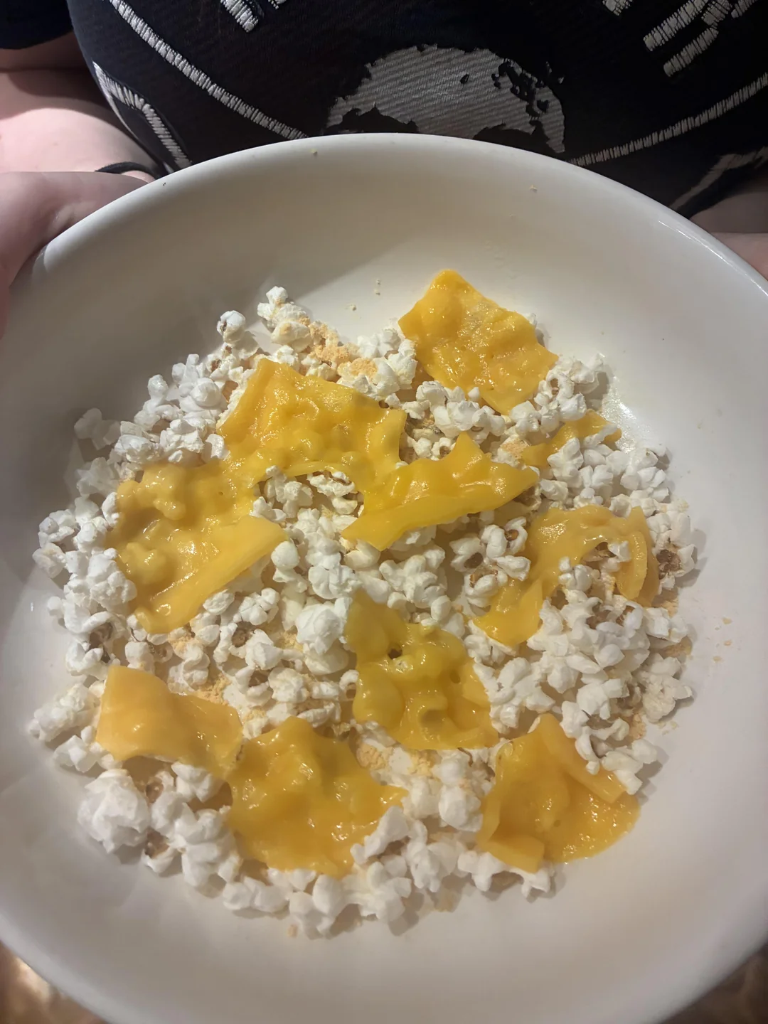 A white bowl filled with popcorn, topped with several pieces of melted yellow cheese. The background shows part of a black shirt with a white graphic design.