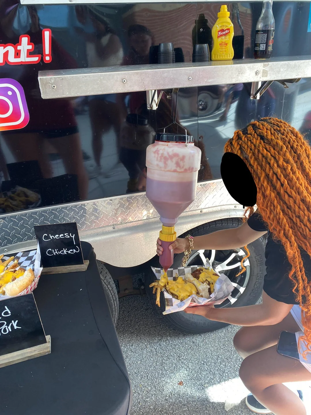 A person with orange braids dispenses ketchup from a large container onto cheesy fries at a food truck, with condiments and menu signs visible nearby.