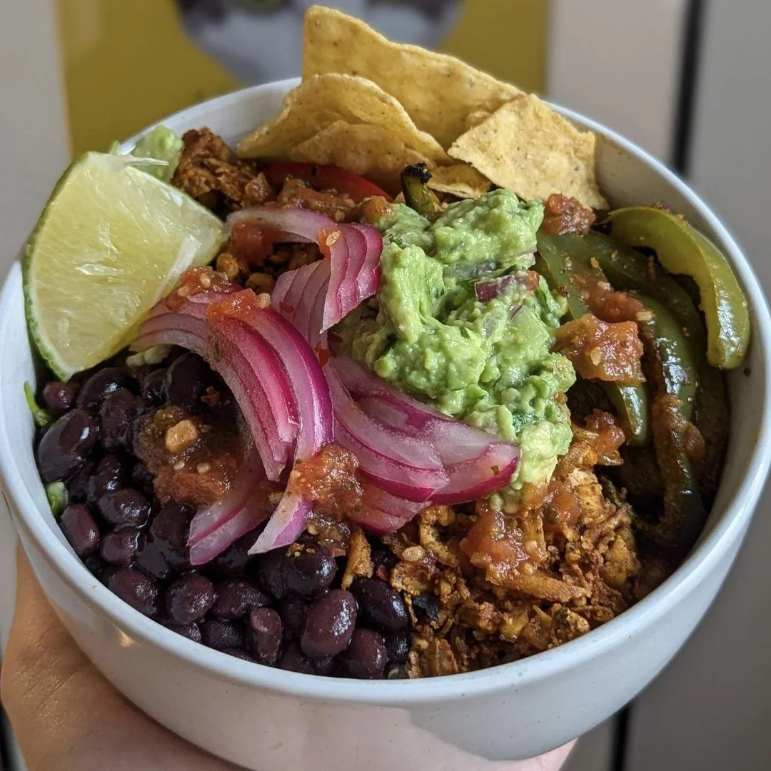 A bowl filled with black beans, shredded chicken, guacamole, pickled red onions, salsa, grilled peppers, tortilla chips, and a lime wedge.