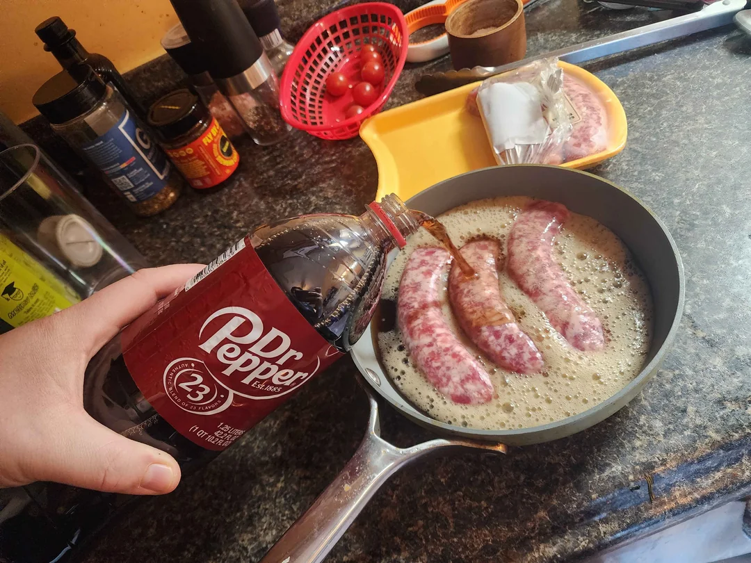 A hand pours Dr. Pepper soda into a frying pan with three sausages cooking on a stove, surrounded by kitchen items and food on a countertop.