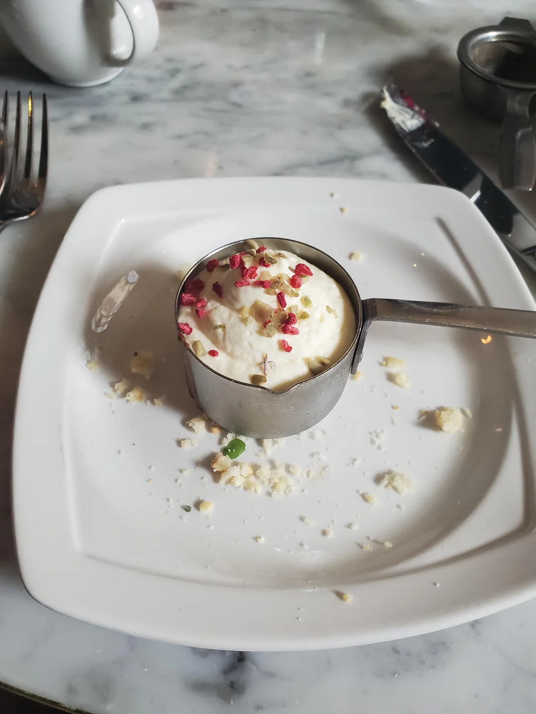 A metal measuring cup filled with vanilla ice cream topped with pink sprinkles sits on a white plate with crumbs, on a marble table with a fork, knife, and cup nearby.
