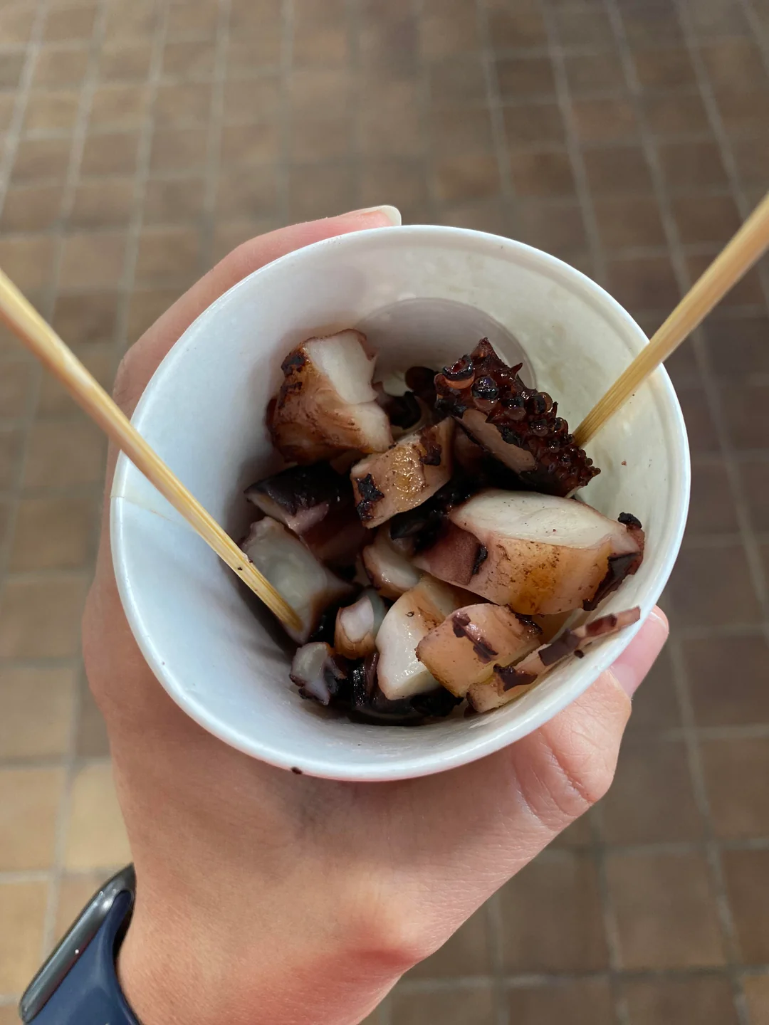 A hand holds a white cup filled with pieces of cooked octopus, skewered with two wooden sticks, over a tiled floor background.