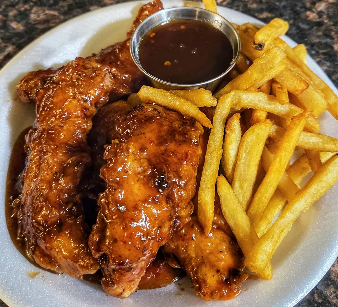 Plate of saucy chicken tenders with a side of golden French fries and a small metal cup of dipping sauce on a white plate.