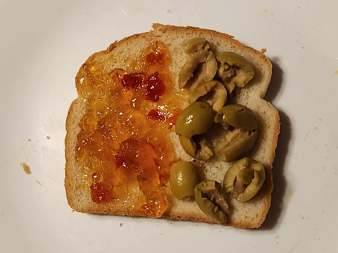 A slice of bread with orange jelly spread on one half and sliced green olives arranged on the other half, on a white plate.