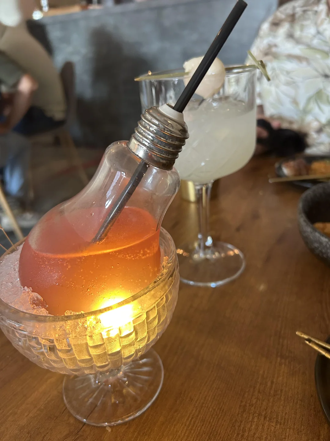 A pink drink served in a light bulb-shaped glass sits on crushed ice, glowing from a light below. In the background, a clear cocktail with a lychee garnish is in a stemmed glass.