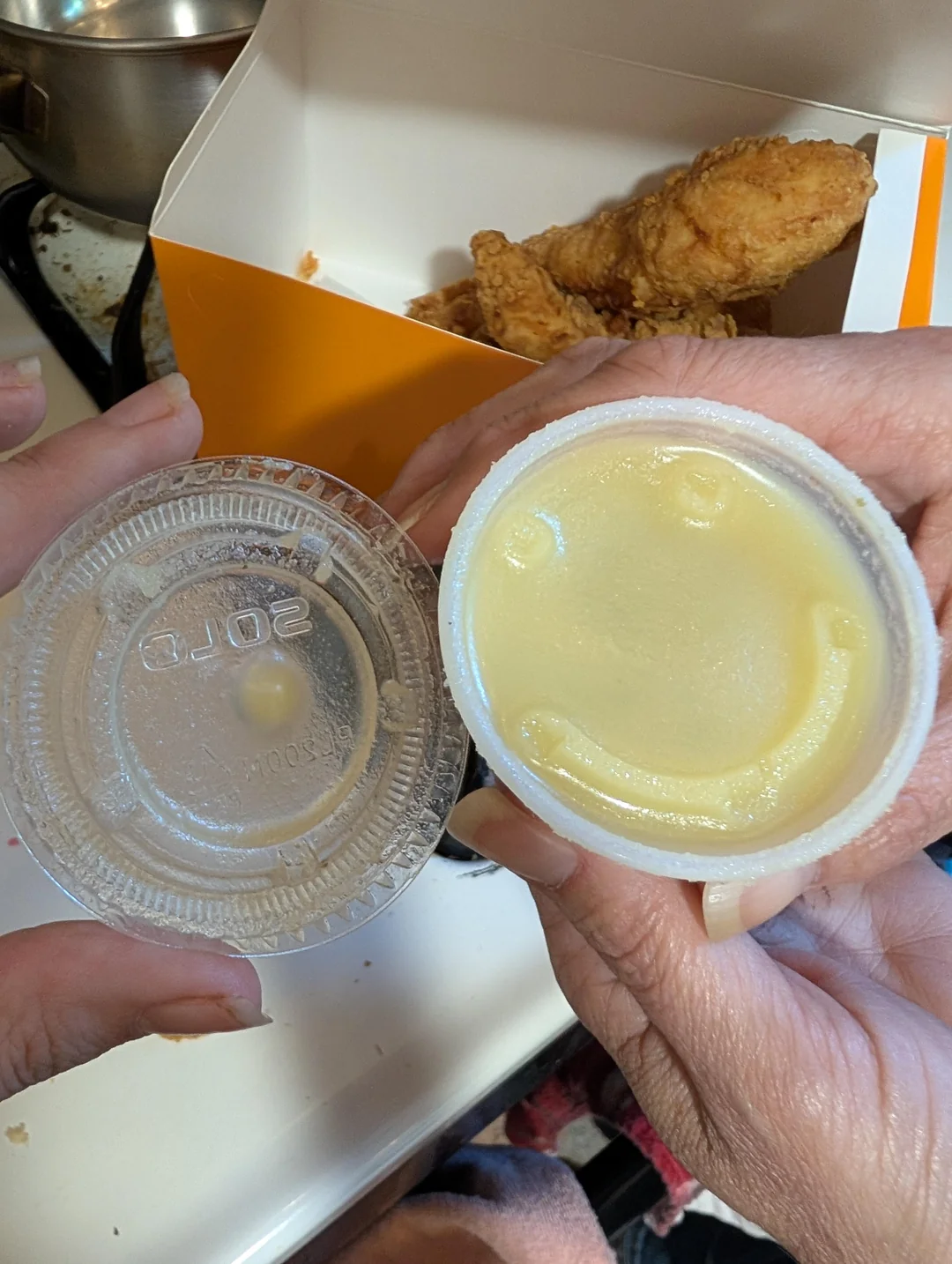A person holds a plastic cup of mashed potatoes with a smiley face imprint in the potatoes. Fried chicken and a takeout box are visible in the background.