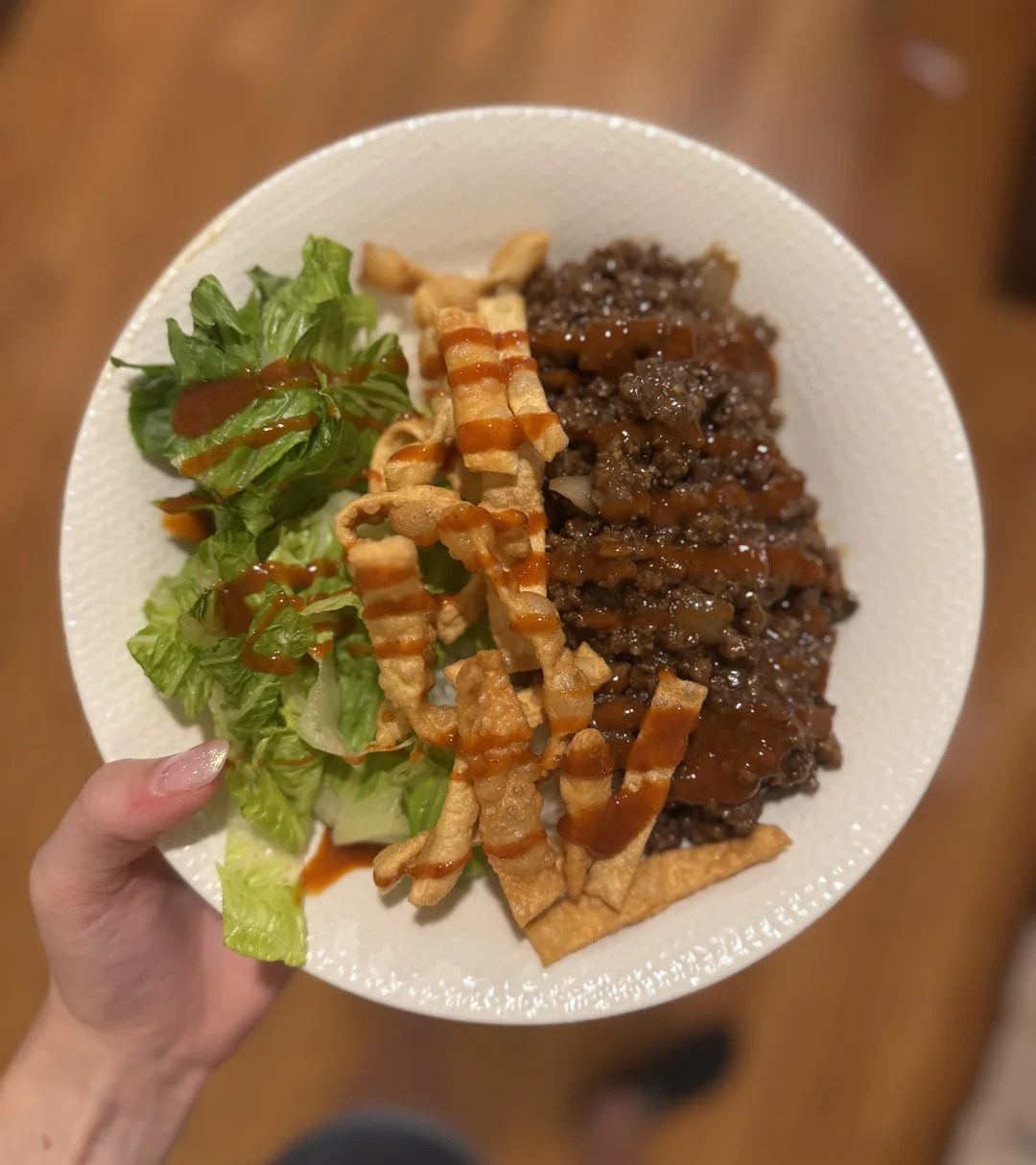 A hand holds a plate with chopped lettuce, crispy wonton strips, and ground beef topped with sauce, on a blurred brown background.
