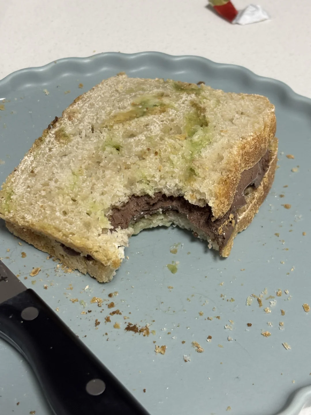 A sandwich with a bite taken out sits on a light blue plate next to a black-handled knife. The filling appears to be chocolate spread and possibly avocado. Crumbs are scattered on the plate.