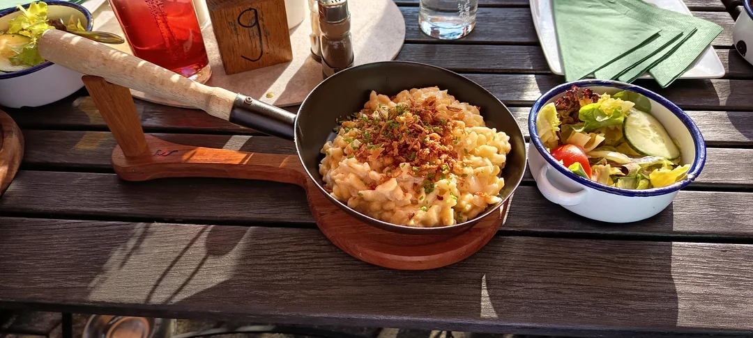 A skillet of macaroni and cheese topped with crispy onions sits on a wooden table next to a bowl of mixed salad, a drink, green napkins, and utensils, all in bright natural sunlight.