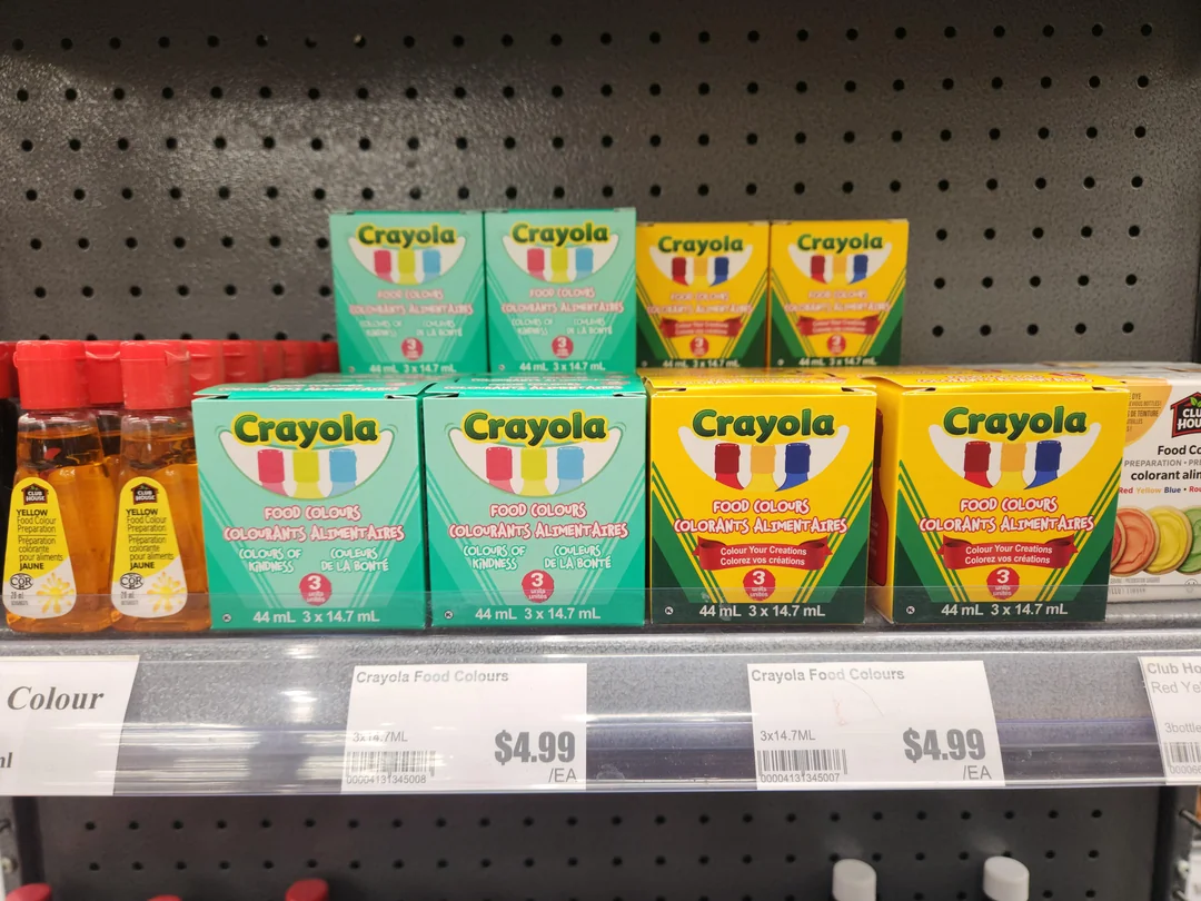 Boxes of Crayola food colors in green and yellow packaging are displayed on a store shelf, priced at $4.99 each. Small bottles with red caps are also partially visible to the left.