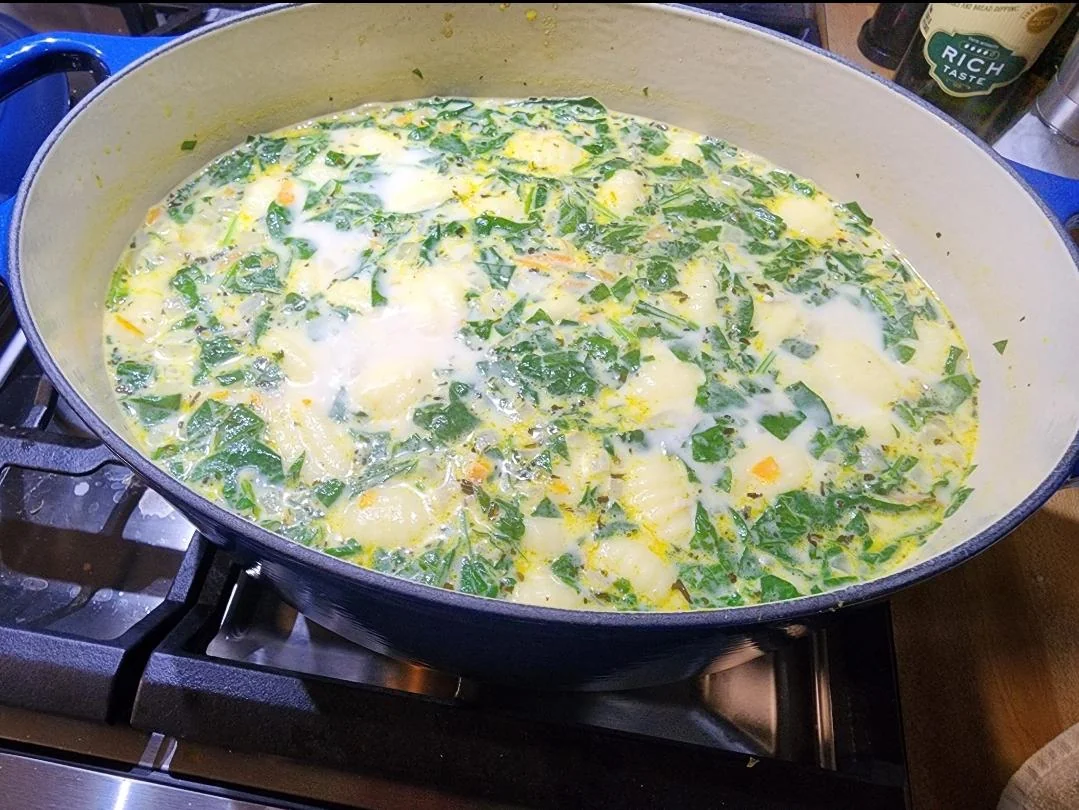 A large pot of creamy soup with leafy greens and potato slices simmers on a stovetop. A bottle labeled “Rich Mate” is partially visible in the background.