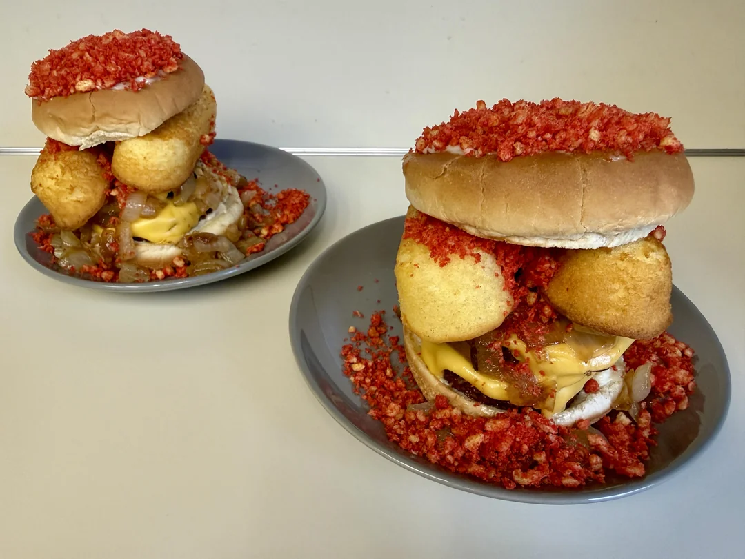 Two burgers topped with cheese, fried appetizers, and a generous amount of crushed red chips sit on gray plates. Crushed chips also surround the burgers on the plates.