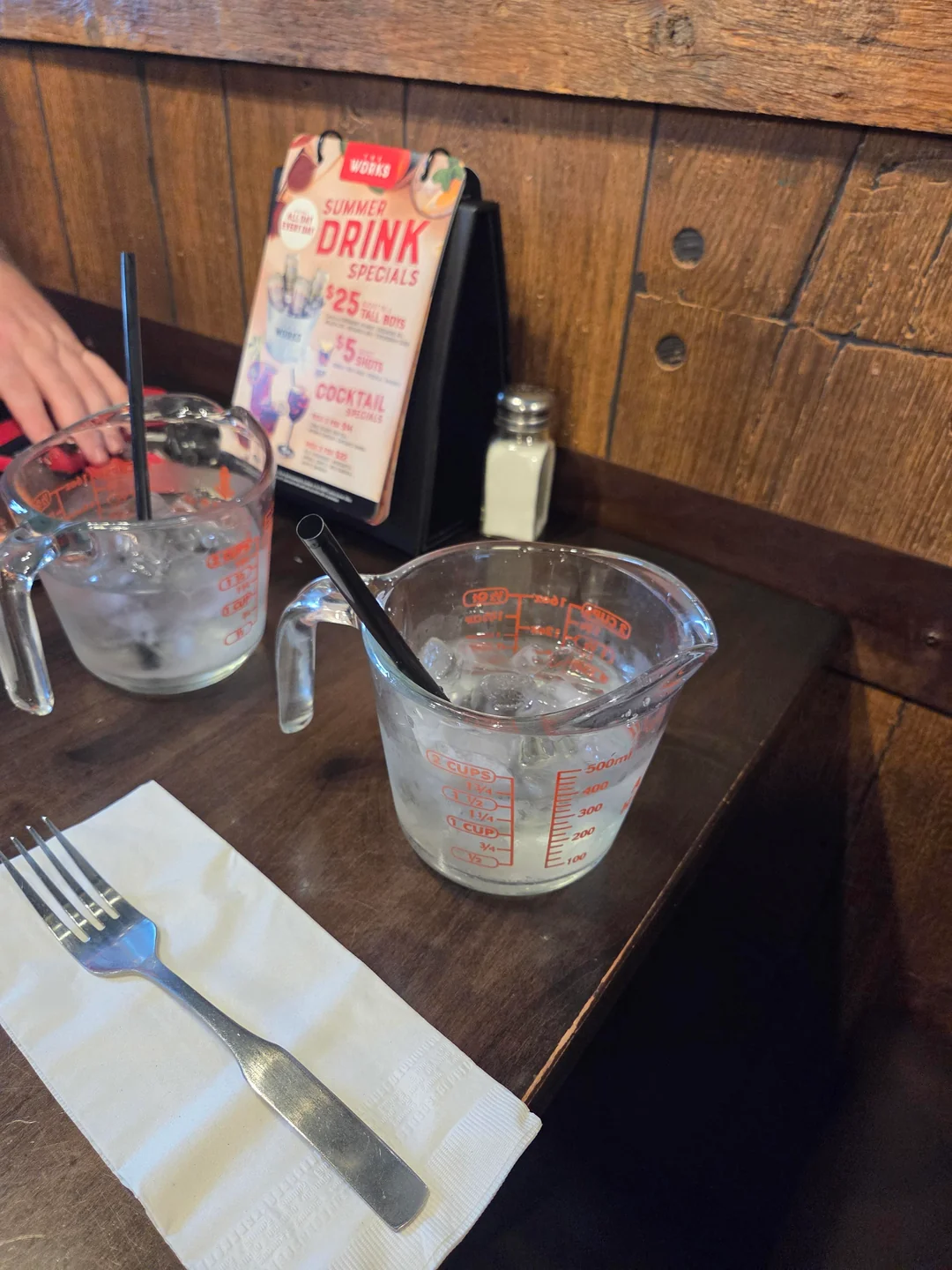 Two measuring cups filled with ice water and straws sit on a dark wooden table next to a fork, napkin, and a drink specials menu in a restaurant setting.