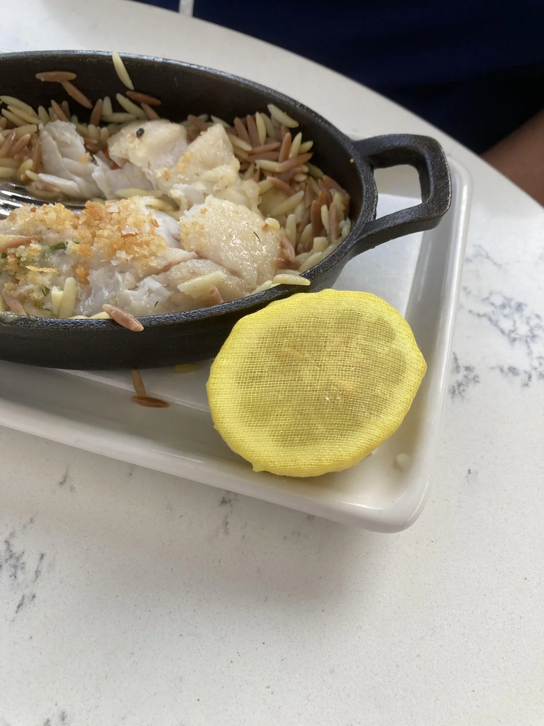 A small skillet with fish and orzo on a white plate, next to a slice of lemon wrapped in yellow mesh. The dish is set on a white marble table.