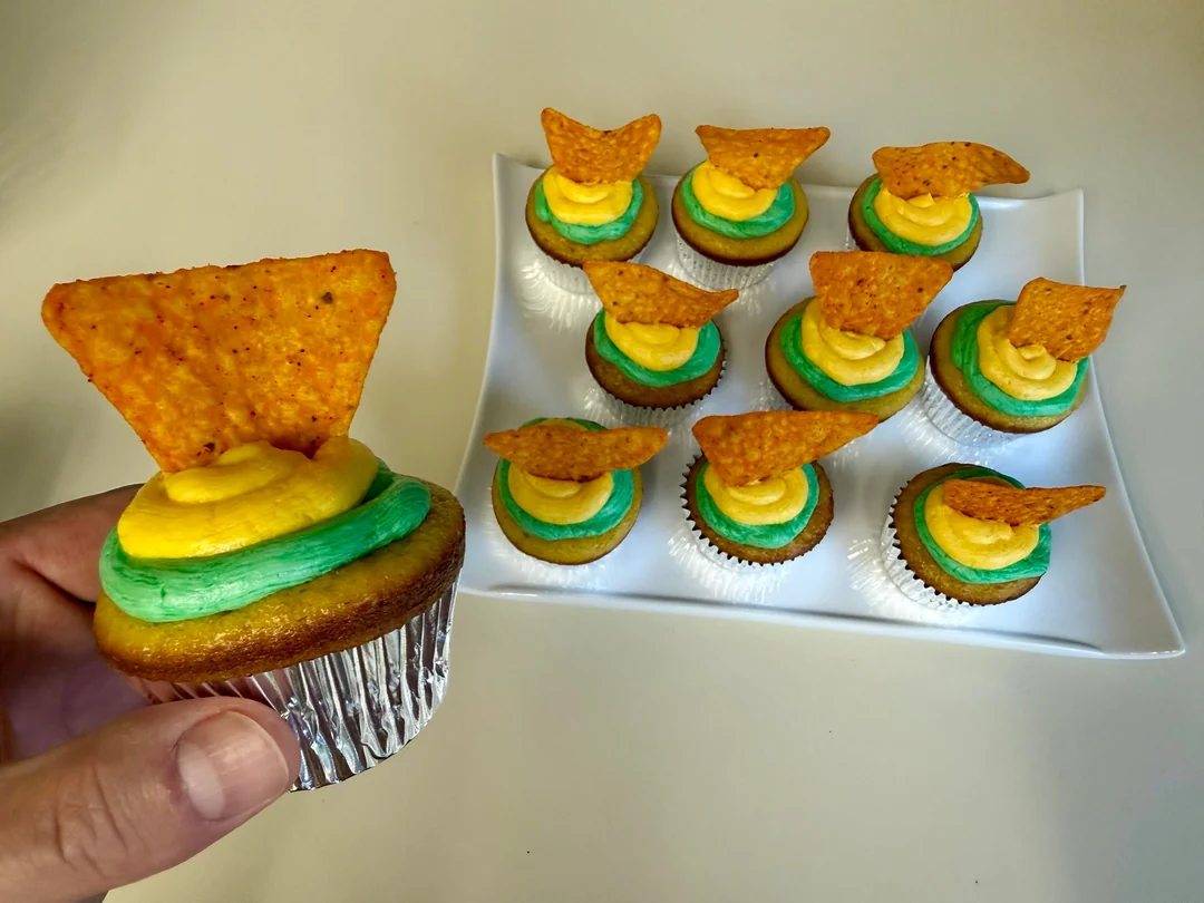 A hand holds a cupcake with yellow and green frosting, topped with a tortilla chip. More similarly decorated cupcakes sit on a white square plate in the background.